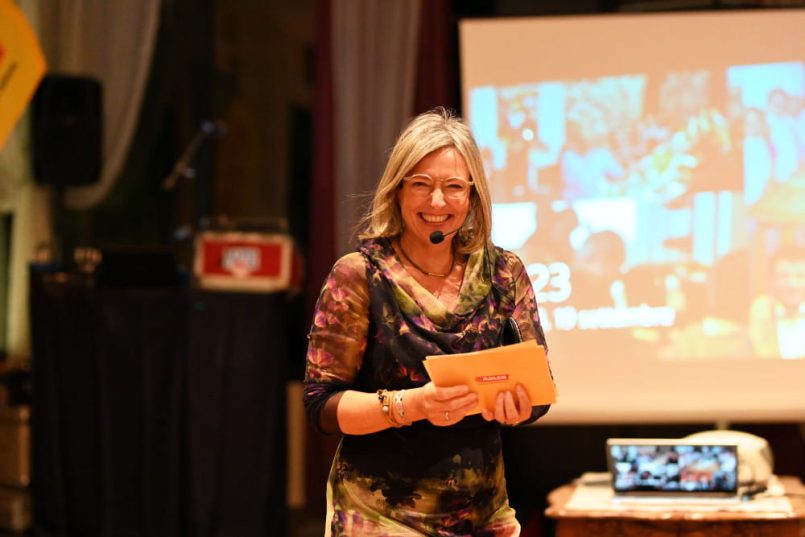 A woman with a microphone and moderation cards smiles into the camera.