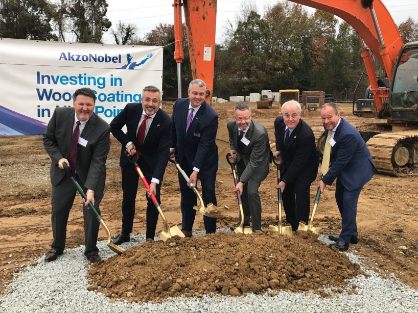 Six men in suits hold golden spades in their hands on a construction site in front of a heap of earth.