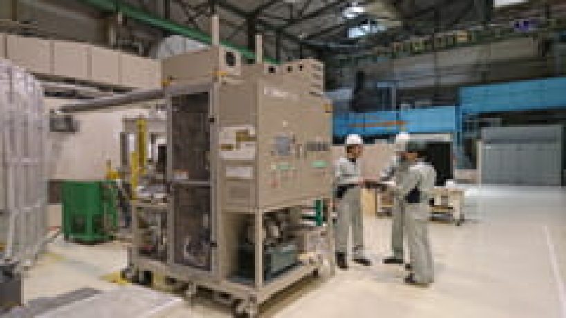 Three men in protective clothing and helmets stand next to a production plant.