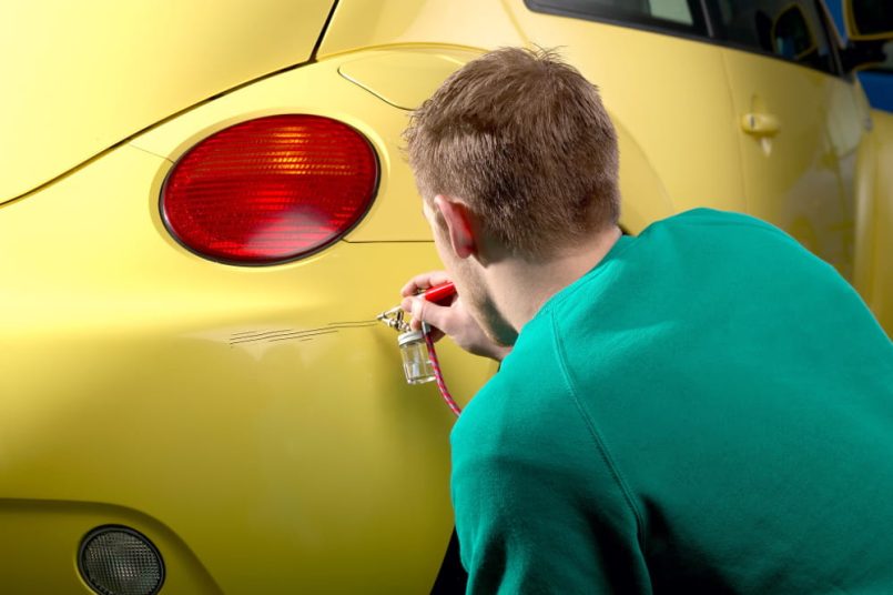 Man kneels in front of a yellow car and fixes a paint defect.