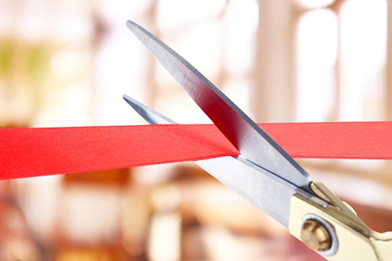 A close-up shows a pair of scissors cutting a red ribbon.