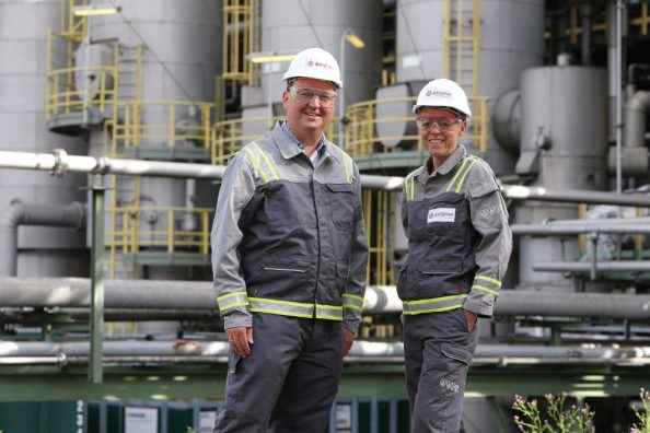 A man and a woman in protective clothing stand in front of a factory.