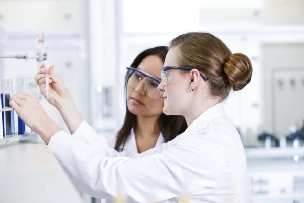 Two women wearing lab coats and goggles look at a test tube.