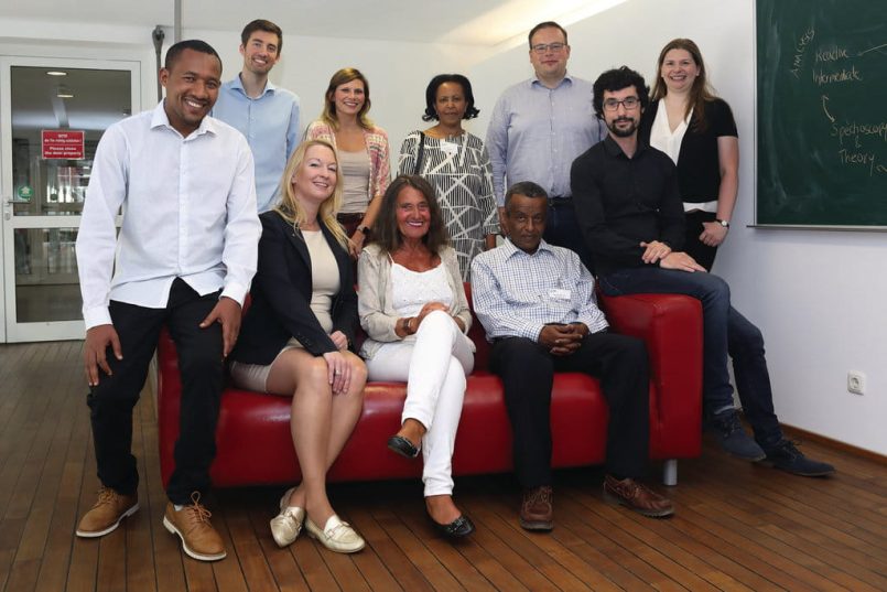 Men and women with light and dark skin colour sit and stand around a red sofa.