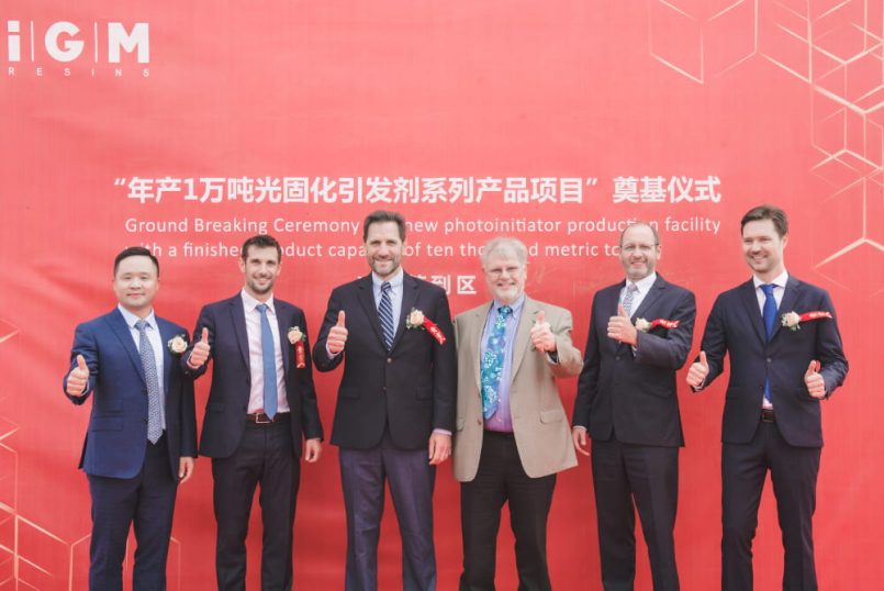 Six men stand next to each other in front of a red display and stretch their thumbs up.