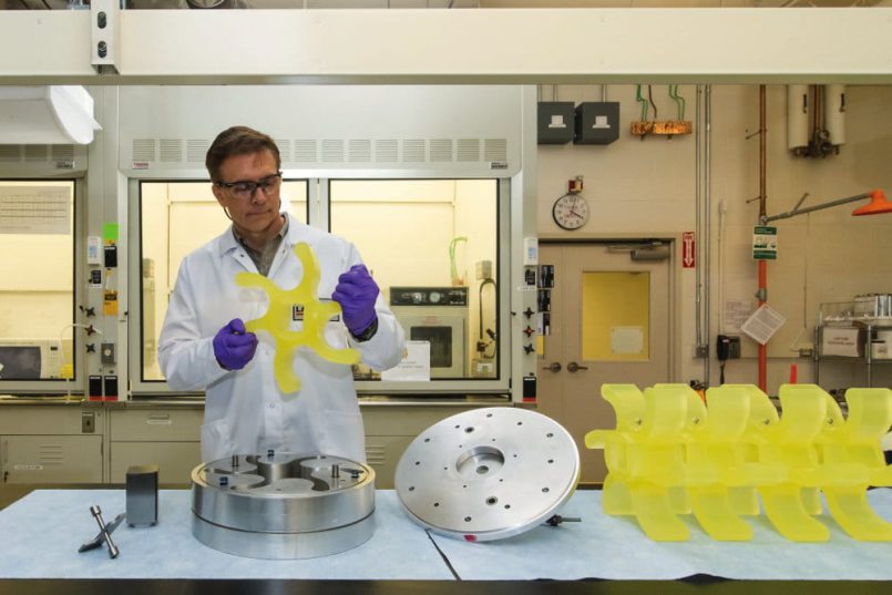 A man in a laboratory with a smock and safety glasses holds a yellow star-shaped wheel in his hands. On the table in front of him is a matching metal mould.