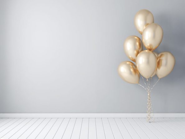 Golden balloons float in the air in front of a white wall and above a white wooden floor.