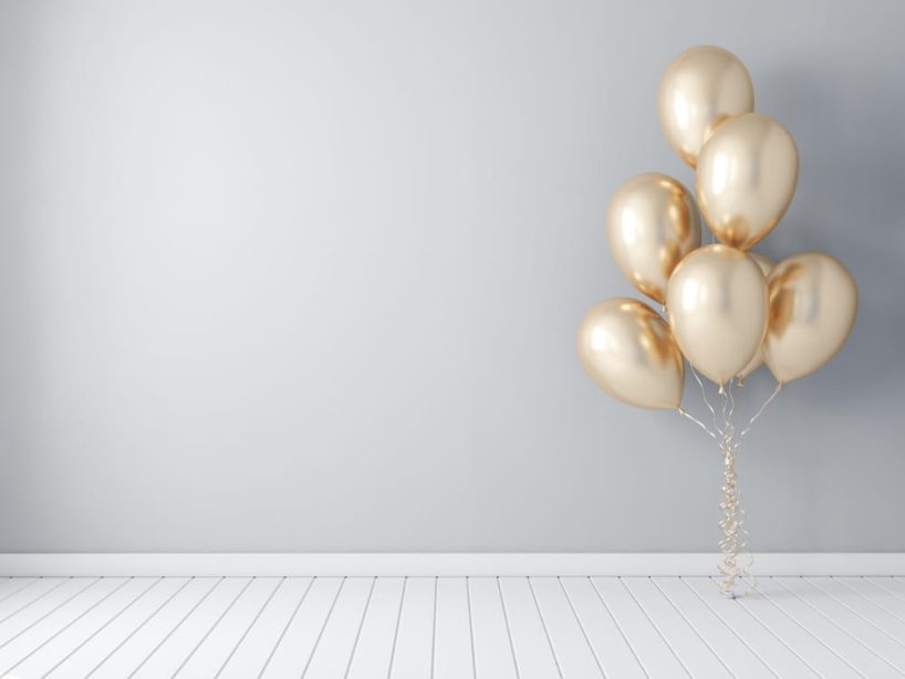 Golden balloons float in the air in front of a white wall and above a white wooden floor.