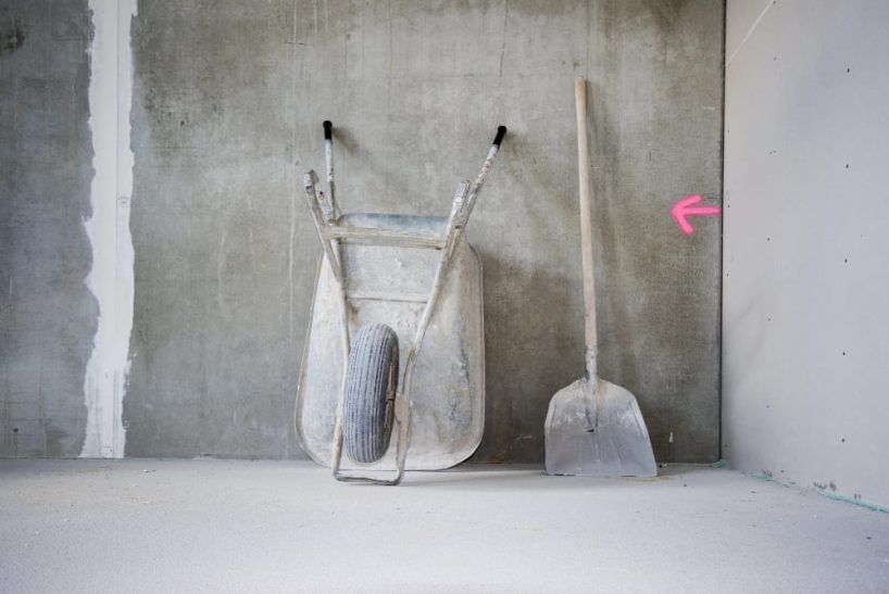 Wheelbarrow and shovel lean next to each other against a wall on a construction site.