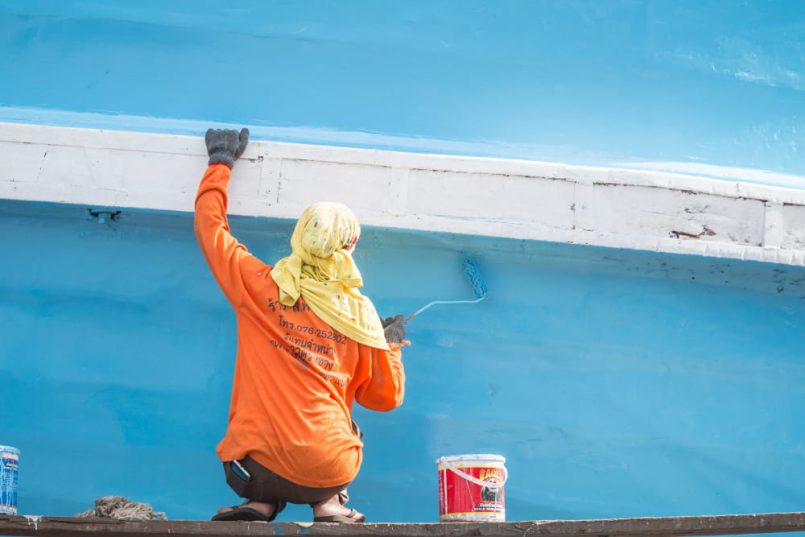 Person in orange clothes and headscarf painting one side of a boat.