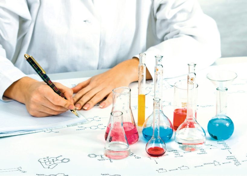 Woman in a white coat sits writing at a table with test tubes on it.