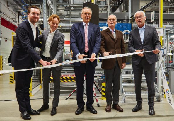 Five men in suits cut a white ribbon in a factory building.