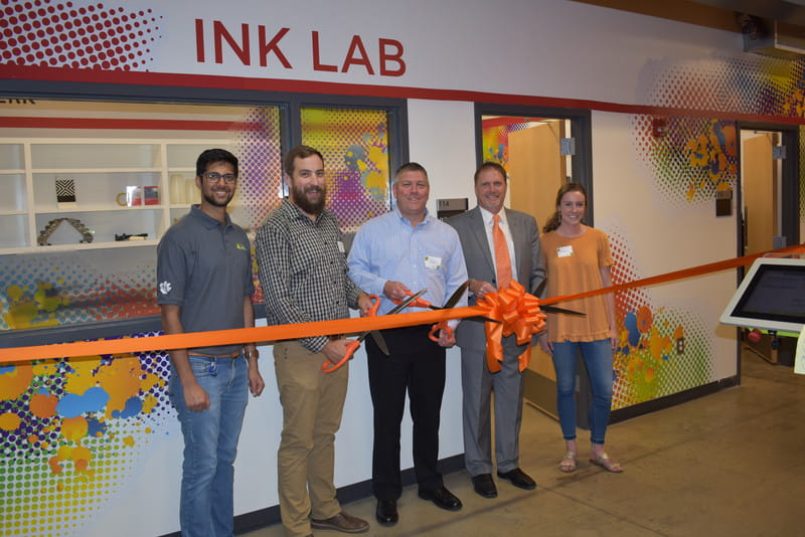 Four men and a woman with big scissors in front of an orange ribbon with bow.